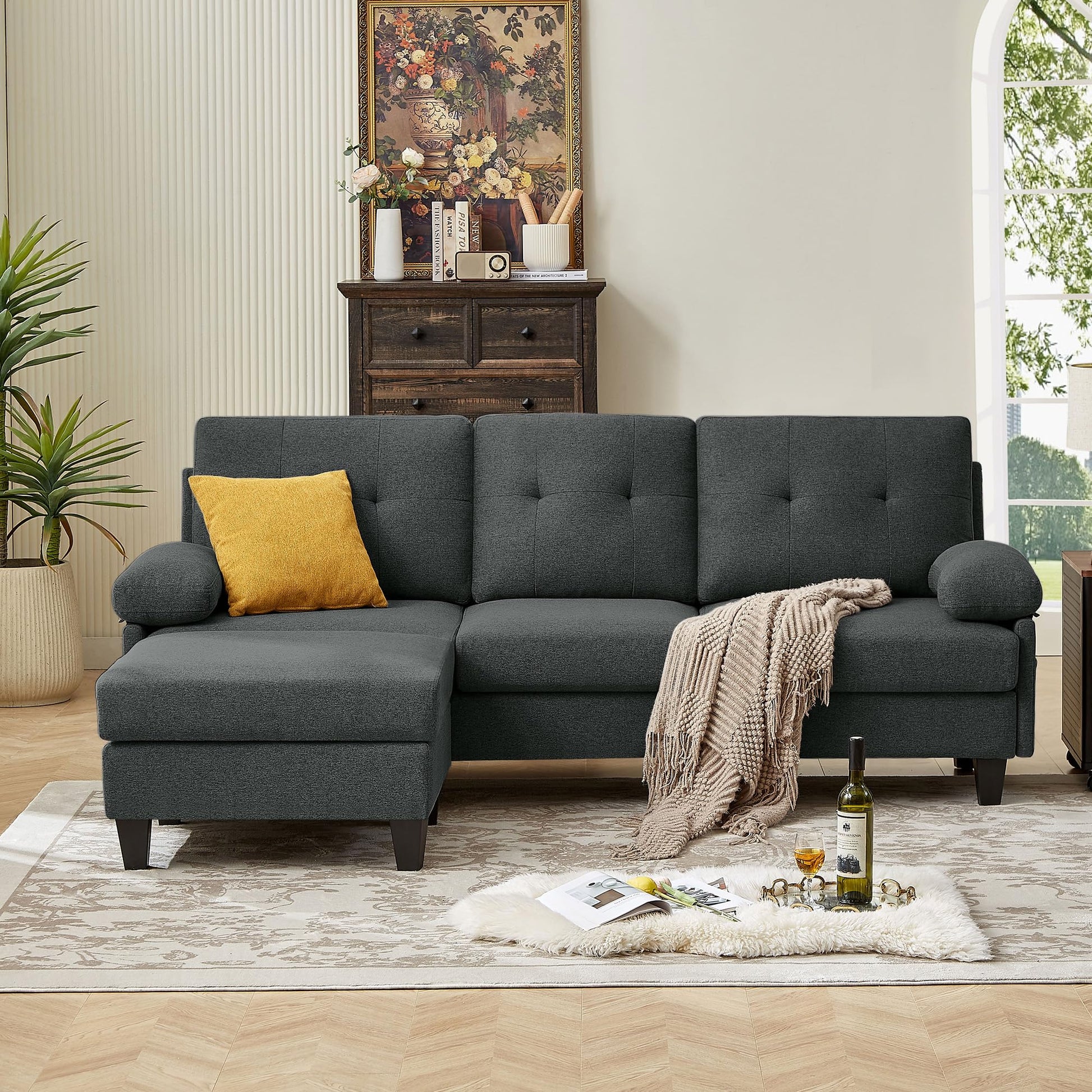 A dark grey convertable sectional sofa with a chaise on the left side, placed in a living room setting with decorative pillows, a rug, and a side table in the background.