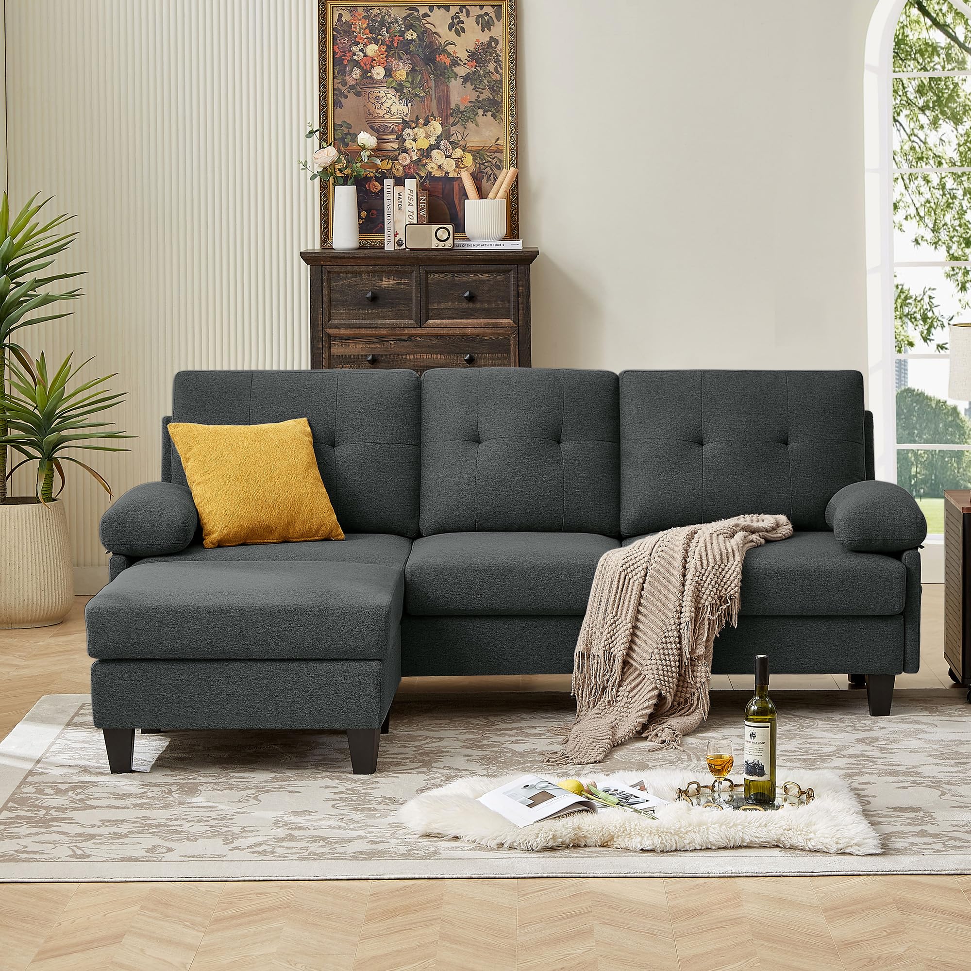 A dark grey convertable sectional sofa with a chaise on the left side, placed in a living room setting with decorative pillows, a rug, and a side table in the background.