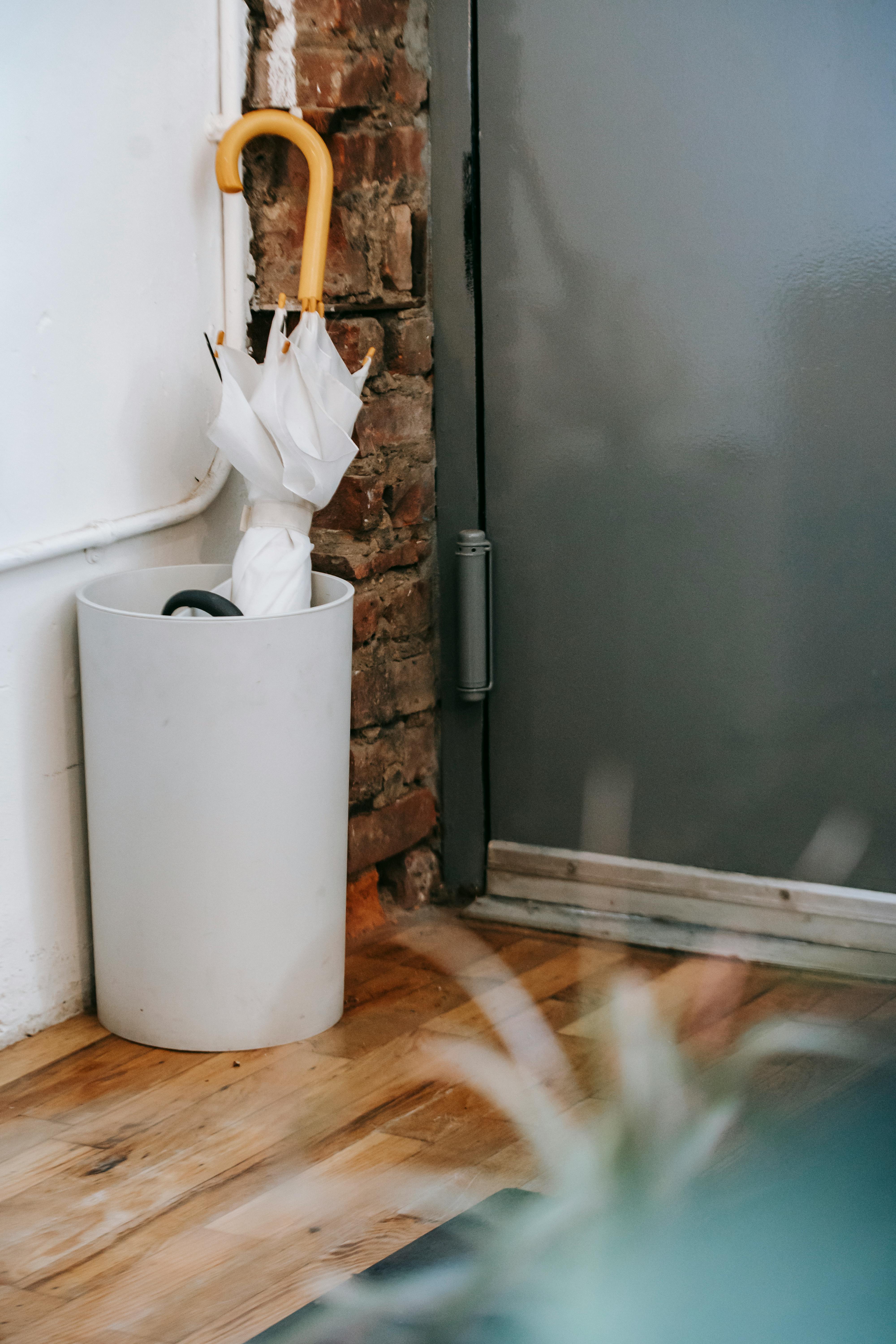 Modern white umbrella stand placed near a doorway, holding umbrellas and plastic bags. A stylish and functional storage solution for any entryway.